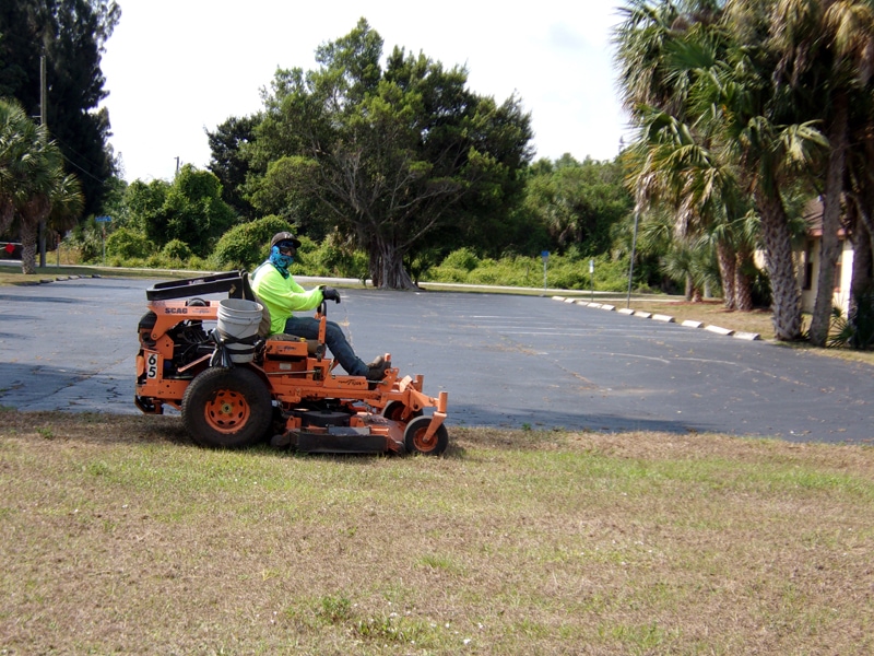 P&T Landscaper on a riding mower with wearing sun-protective clothing and sunglasses while mowing grass and a bucket to pick up trash for Lee County Department of Transportation